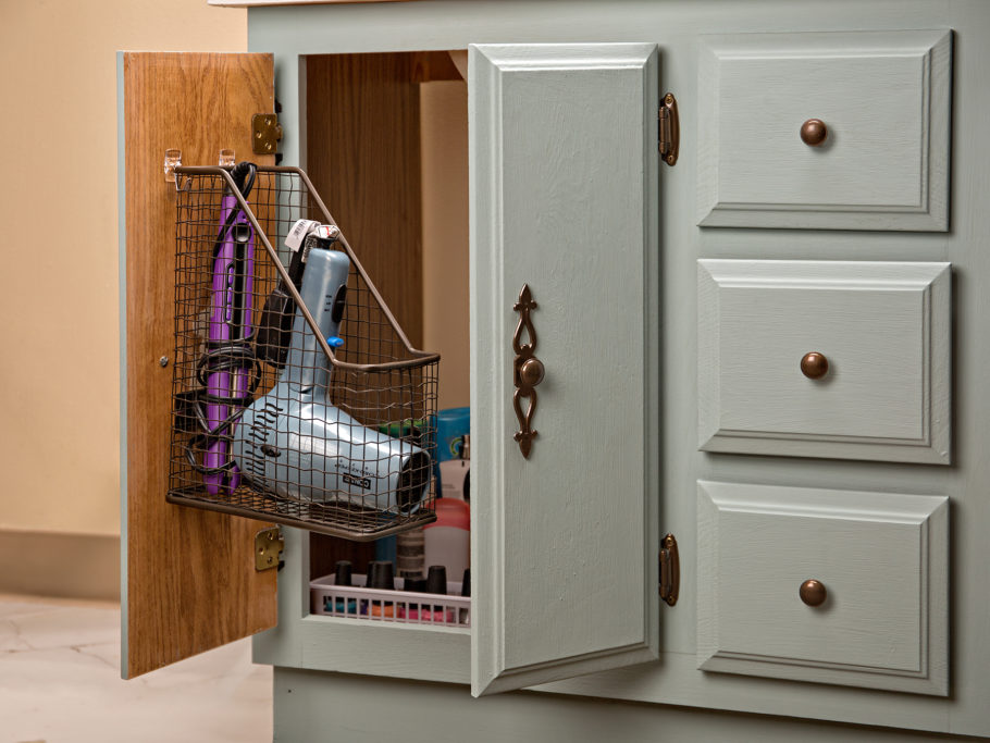 Open cabinet door under bathroom sink with wire binder tray holding hairdryer and flat iron