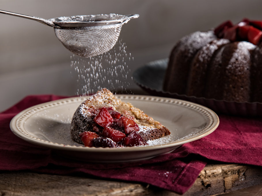 Slice of rye pound cake with plum compote and powdered sugar being sprinkled atop