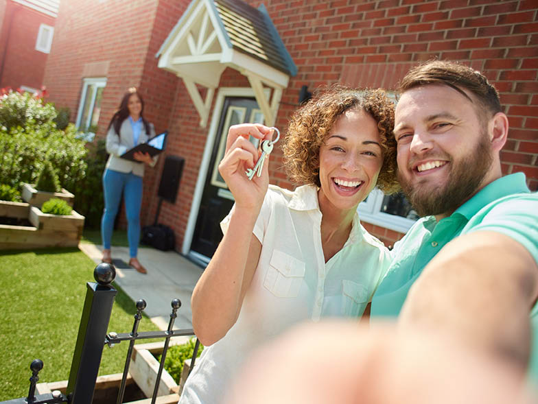 A couple taking a selfie after purchasing a home with their real estate agent in the background