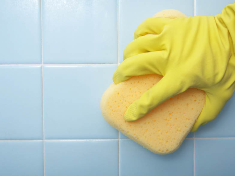 Gloved hand scrubbing tiled bathroom wall with sponge