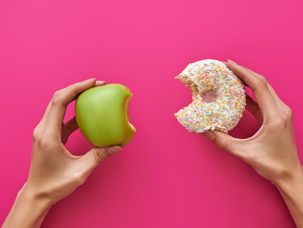 Hand on left holding apple with a bite out of it, hand on right holding donut with a bite out of it