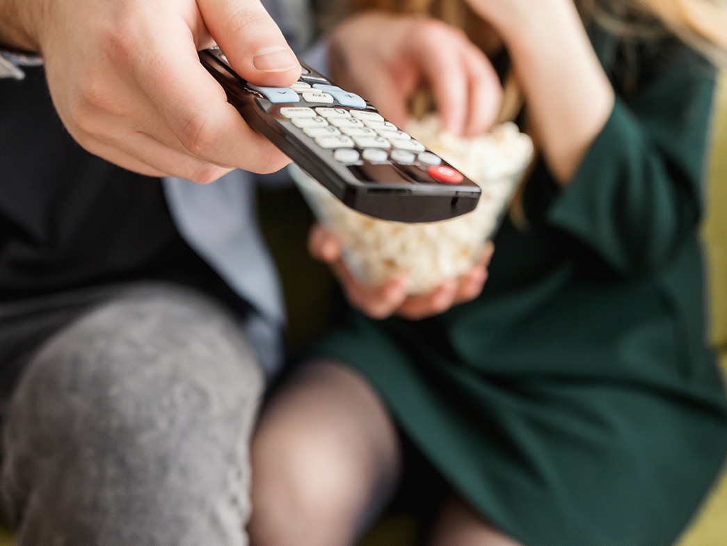 Person holding tv remote up with other person holding popcorn in background