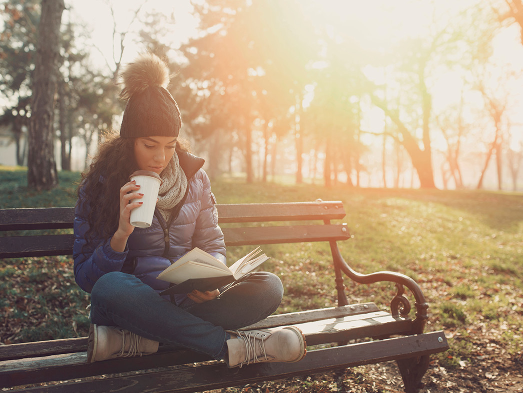 Woman reading book and drinking coffee on park bench