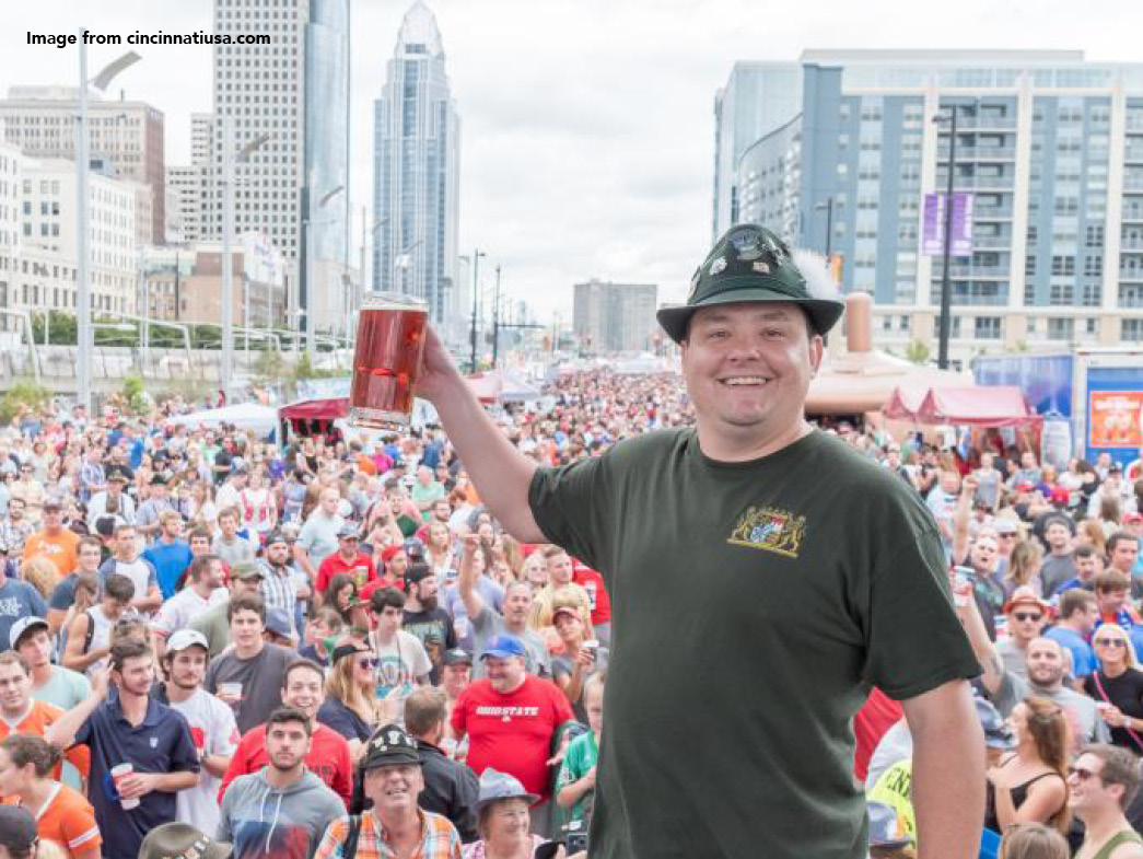 Man smiling and holding up beer in front of crowd. City buildings can be seen in the background.