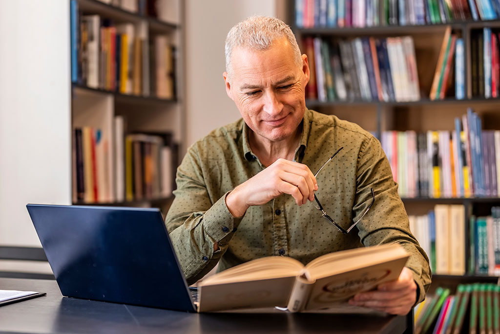 Entertainmment news Man reading book with laptop open in library